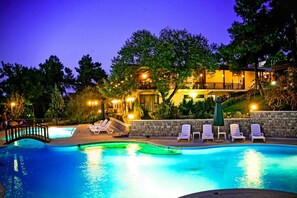 A swimming pool with lounge chairs and a stone wall, surrounded by trees and a building with illuminated windows.