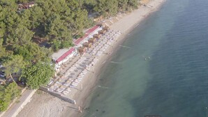 An aerial view of a beach with rows of sun loungers and umbrellas, a building with a red roof, and a clear view of the sea. - Troia Tusan Hotel (Çanakkale)