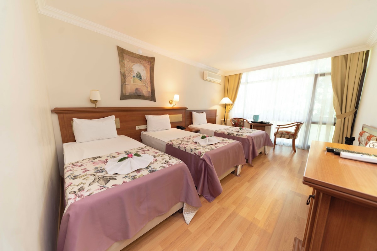 A hotel room with three beds, each with a floral bedspread and white pillows. There is a desk with a lamp, a chair, and a window with curtains.