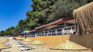 A beachside restaurant with outdoor seating, including sun loungers and umbrellas, and a wooden fence separating the dining area from the beach. - Troia Tusan Hotel (Çanakkale)