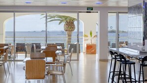 A modern restaurant with a view of the sea, featuring high tables and chairs, a large plant, and a bar area. - Hotel Ibiza Playa (Ibiza Town)