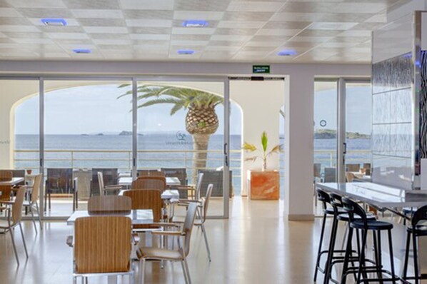 Un restaurante moderno con vistas al mar, que cuenta con mesas y sillas altas, una planta grande y una zona de bar.