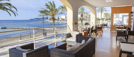 A seaside restaurant with outdoor seating, palm trees, and a view of the sea.