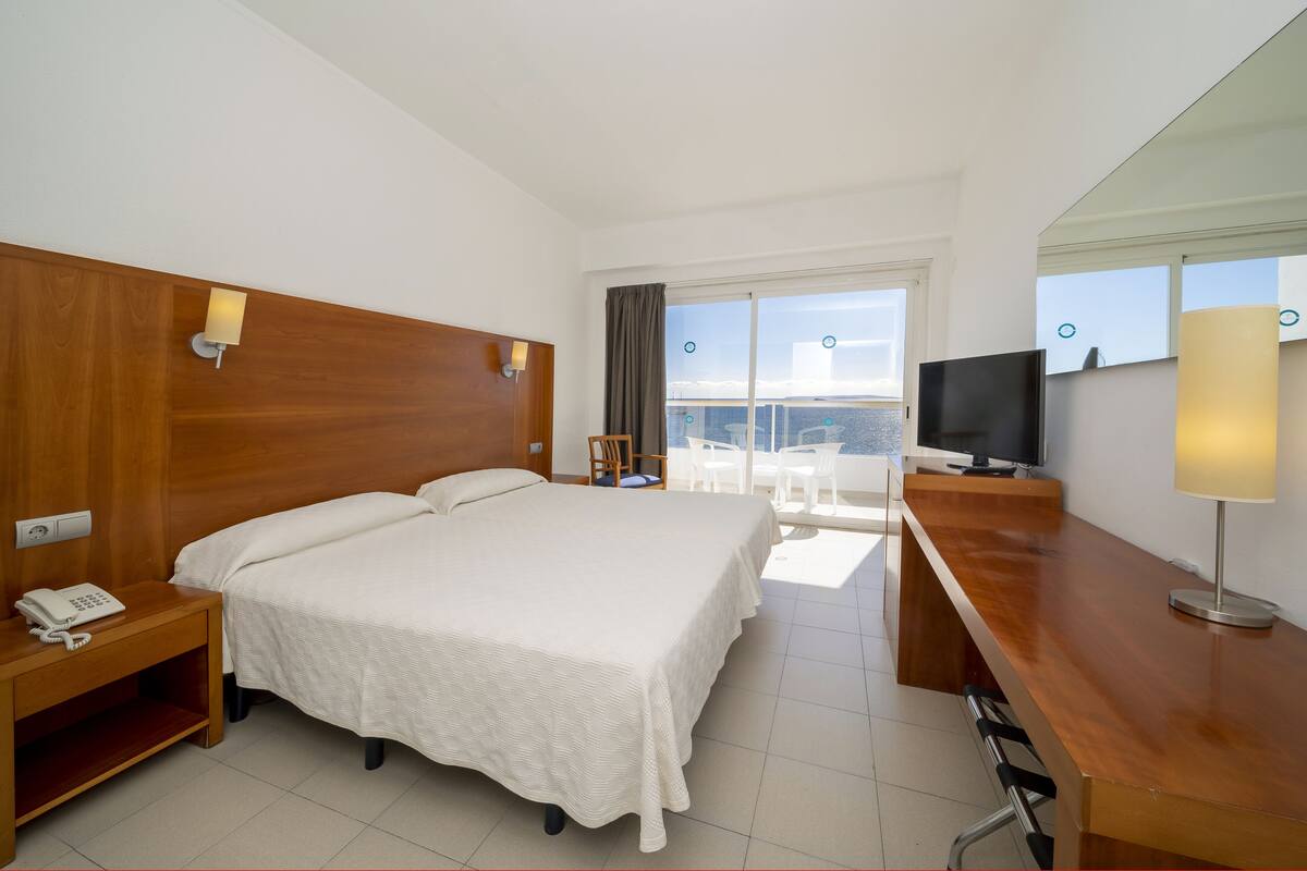 Habitación de hotel con una cama grande, un escritorio con televisor, teléfono y vistas a la ciudad a través de amplios ventanales.