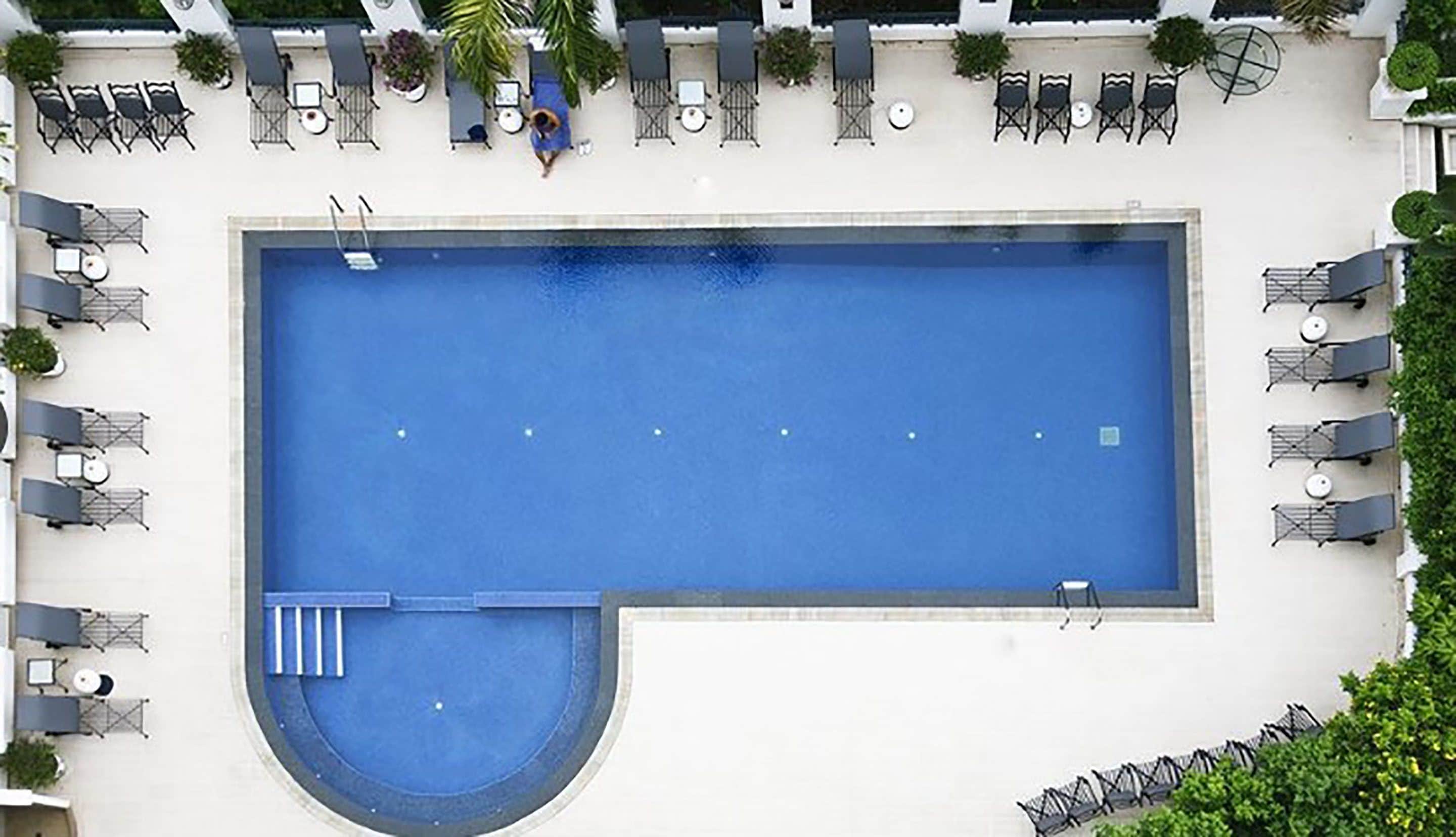 An aerial view of a rectangular swimming pool with a small circular section, surrounded by multiple lounge chairs and tables.