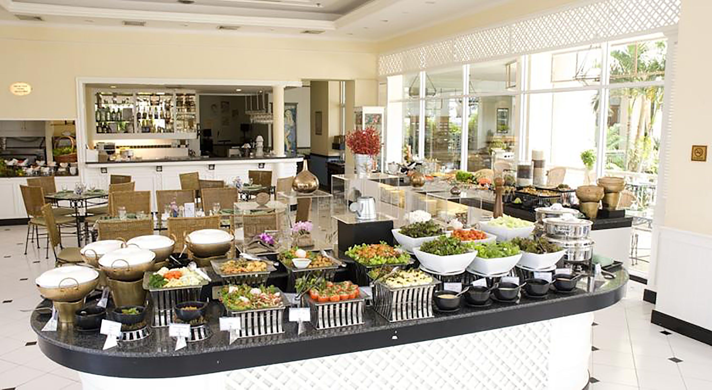 A buffet area with various food stations, a bar in the background, and a view of the outdoors through large windows.