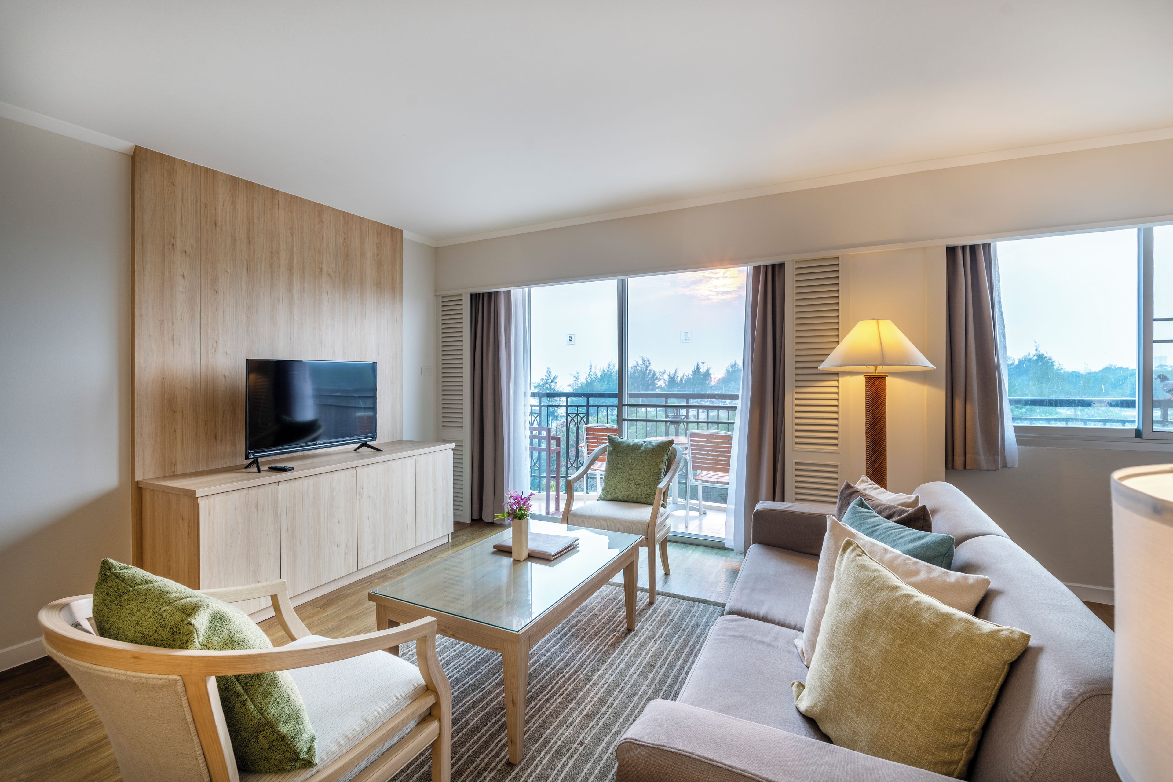 A modern hotel room with a flat-screen TV, a sofa, a chair, a glass coffee table, and a balcony with outdoor furniture.