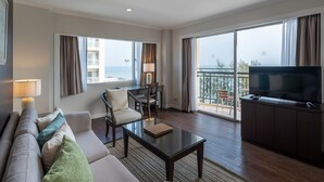 Quarto de hotel moderno com sofá, poltronas, mesa de centro, televisão e varanda com vista para o mar.