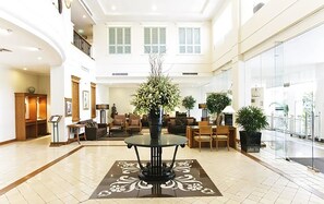 A spacious lobby with a central round table, multiple seating areas, and a large window providing natural light.