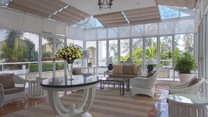 Uma sala de sol com janelas amplas, uma mesa de vidro, móveis de vime e um vaso com planta.