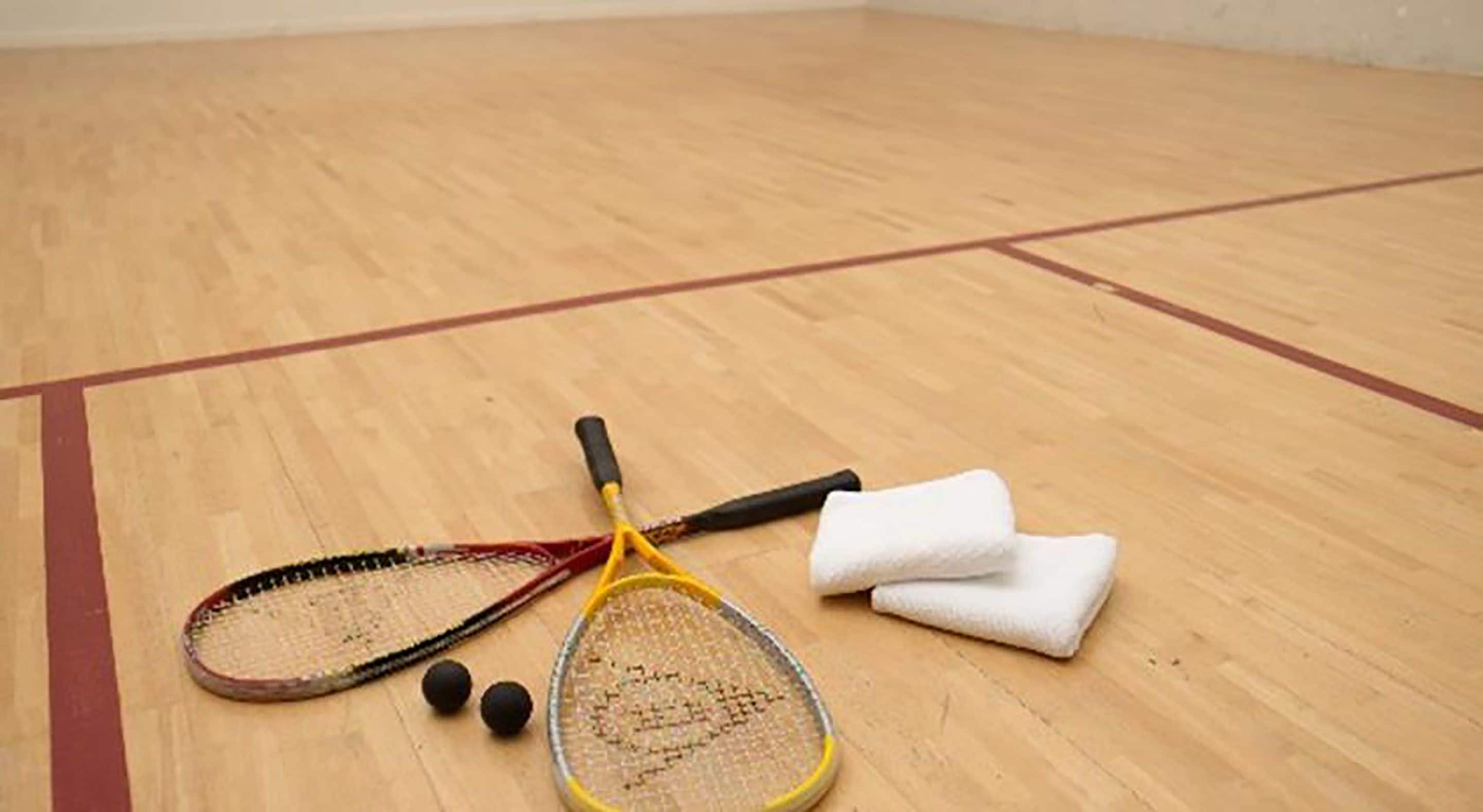 En squashhall med trägolv, ett tennisracket, ett squashracket och två svarta bollar.