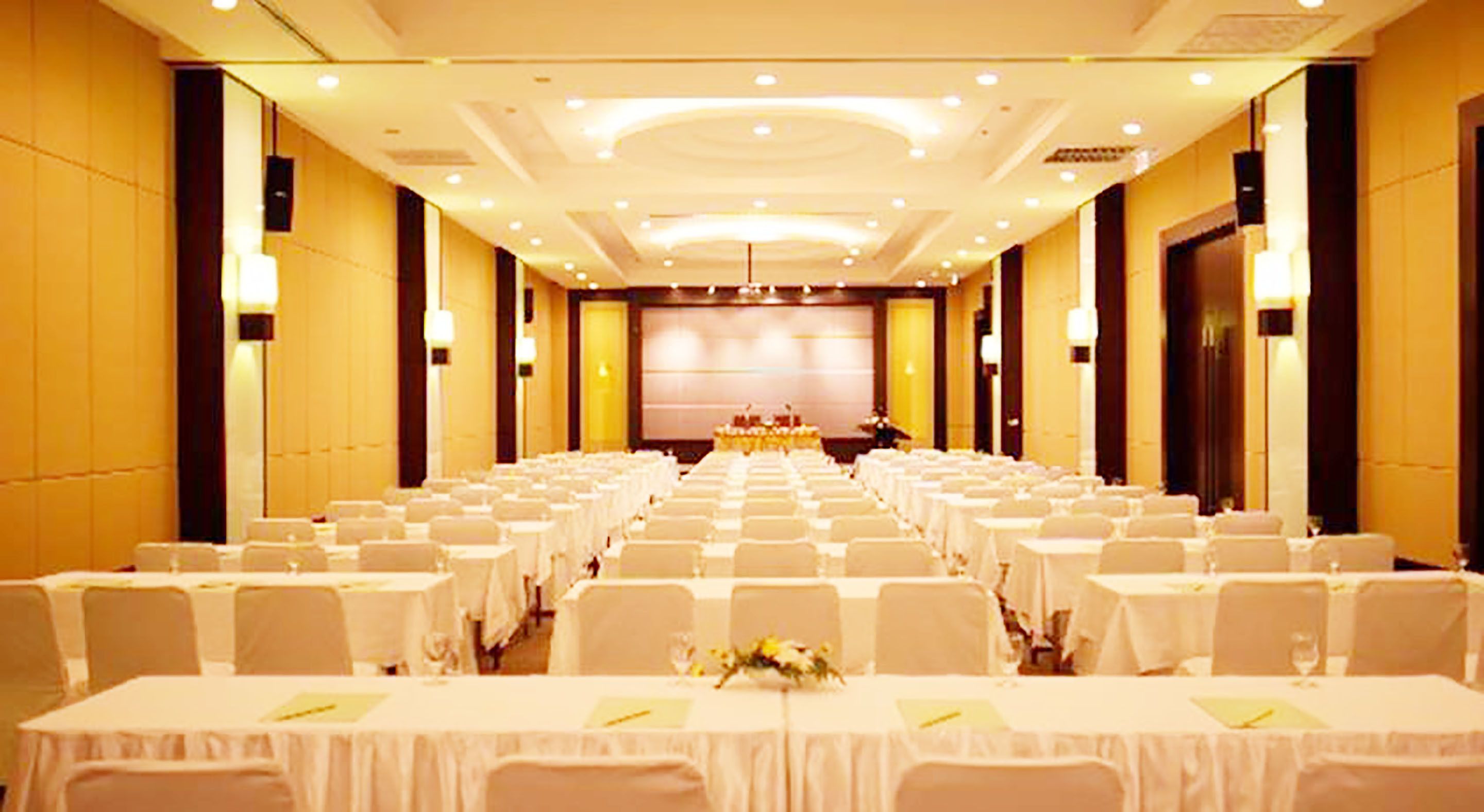 A conference room with rows of tables and chairs, a stage at the front, and a large screen.