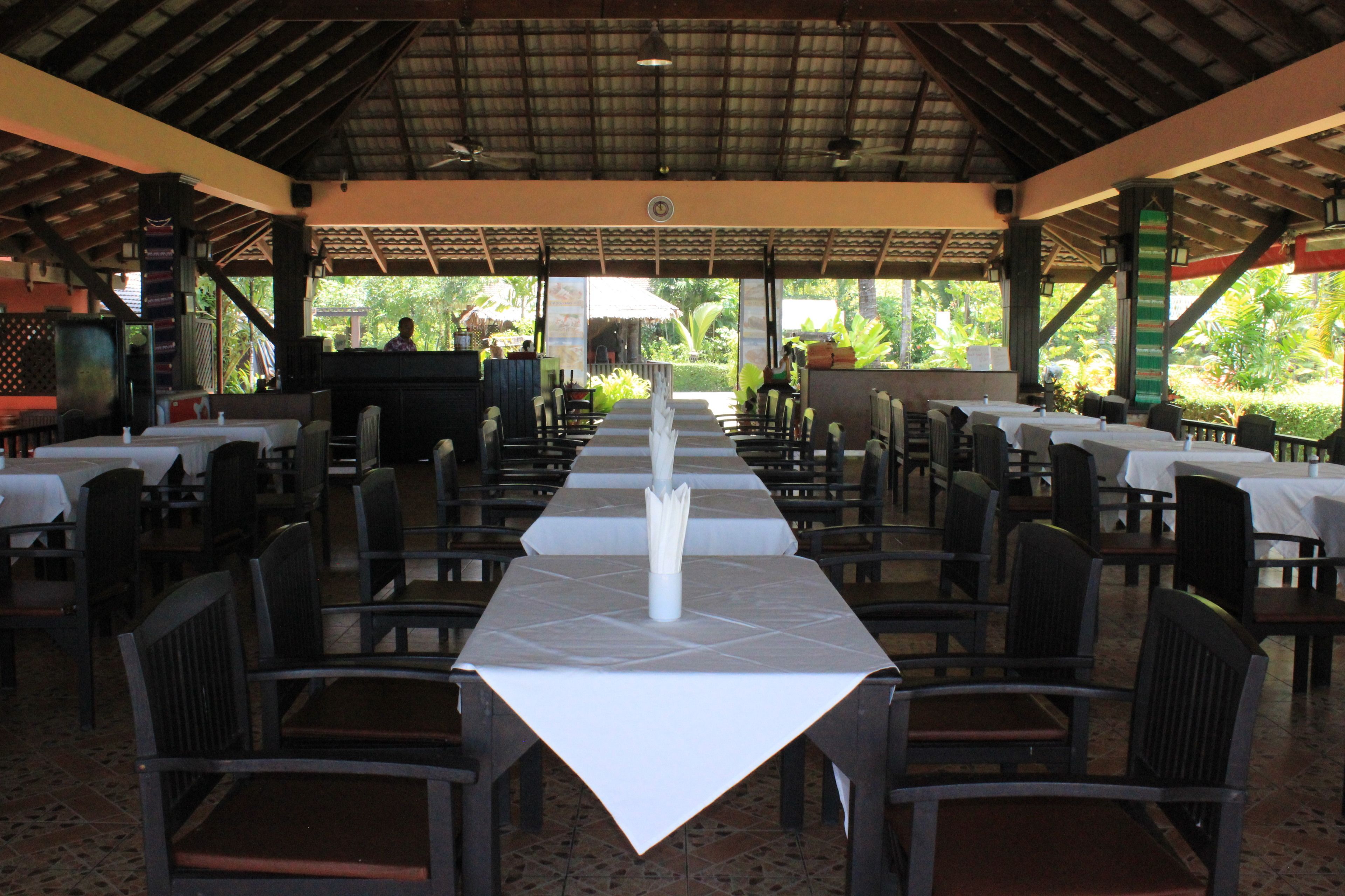 A covered dining area with tables and chairs, a central decorative element, and a view of greenery outside.