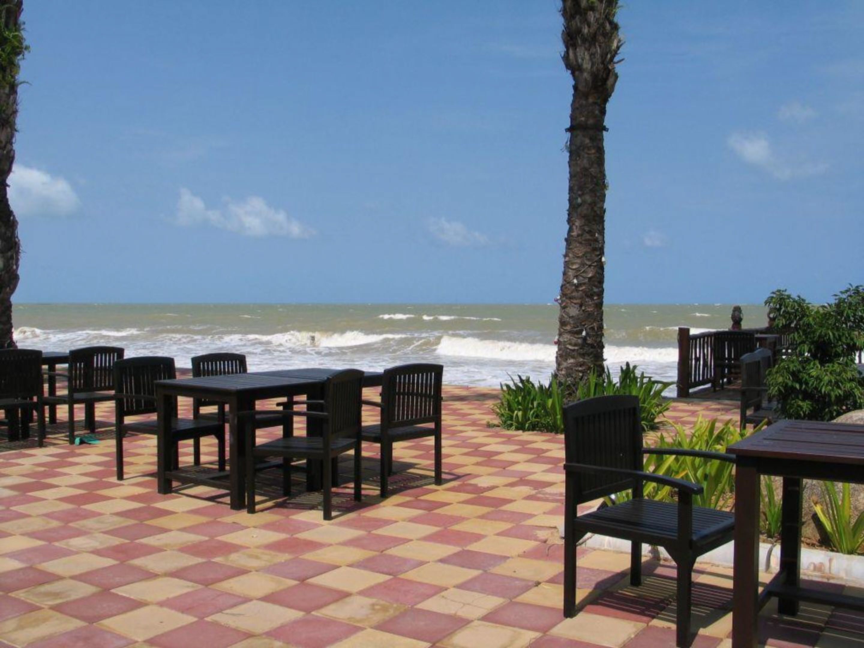 An outdoor seating area with a checkered floor, tables, and chairs, overlooking the ocean with waves crashing onto the shore.