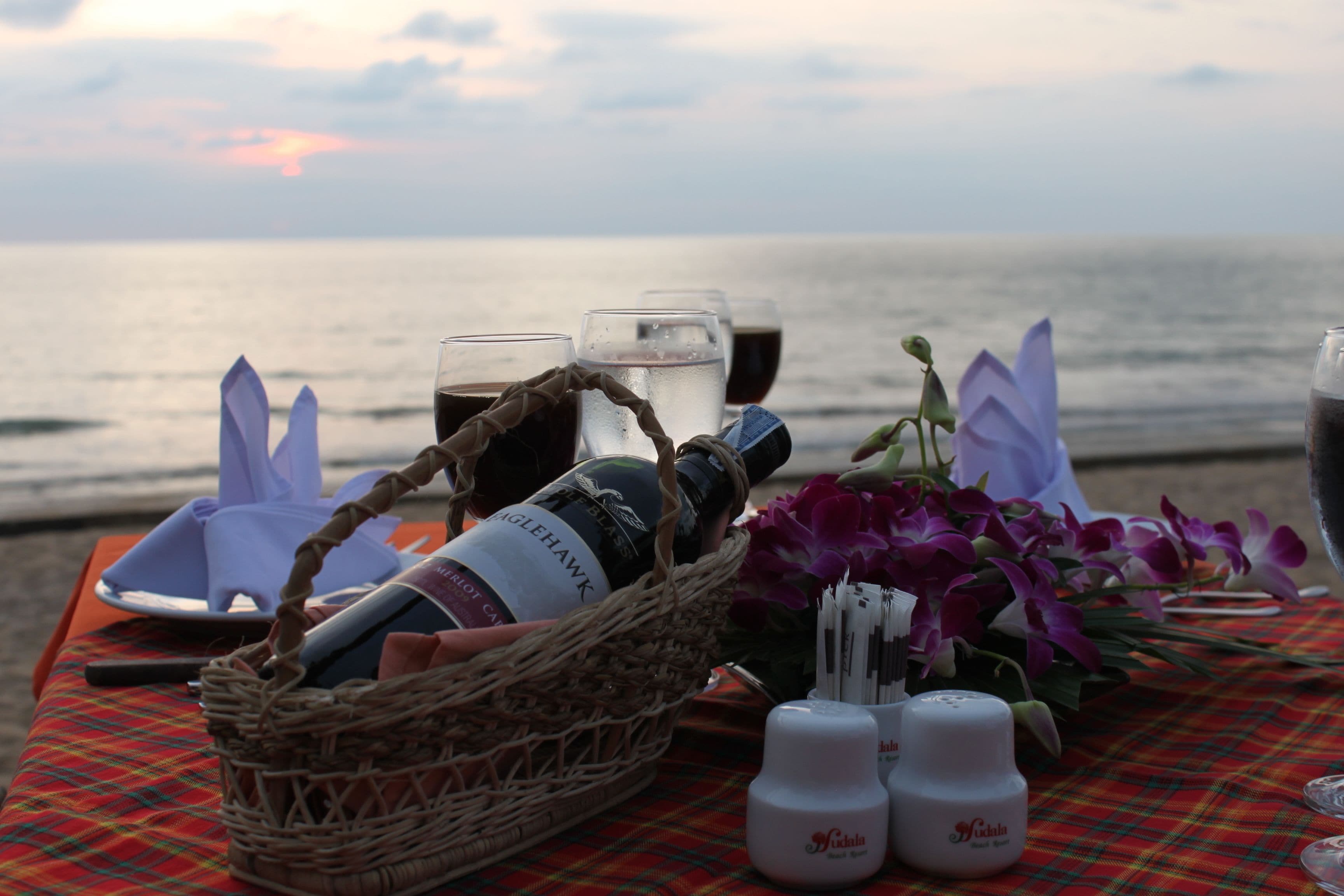 A table set with a bottle, glasses, flowers, and condiments by the ocean.