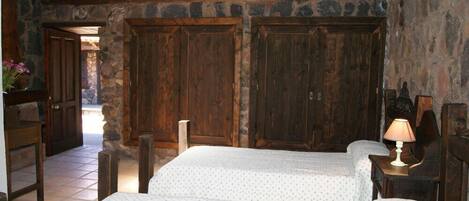 Una habitación rústica con dos camas, puertas de madera, paredes de piedra y suelo de baldosas.