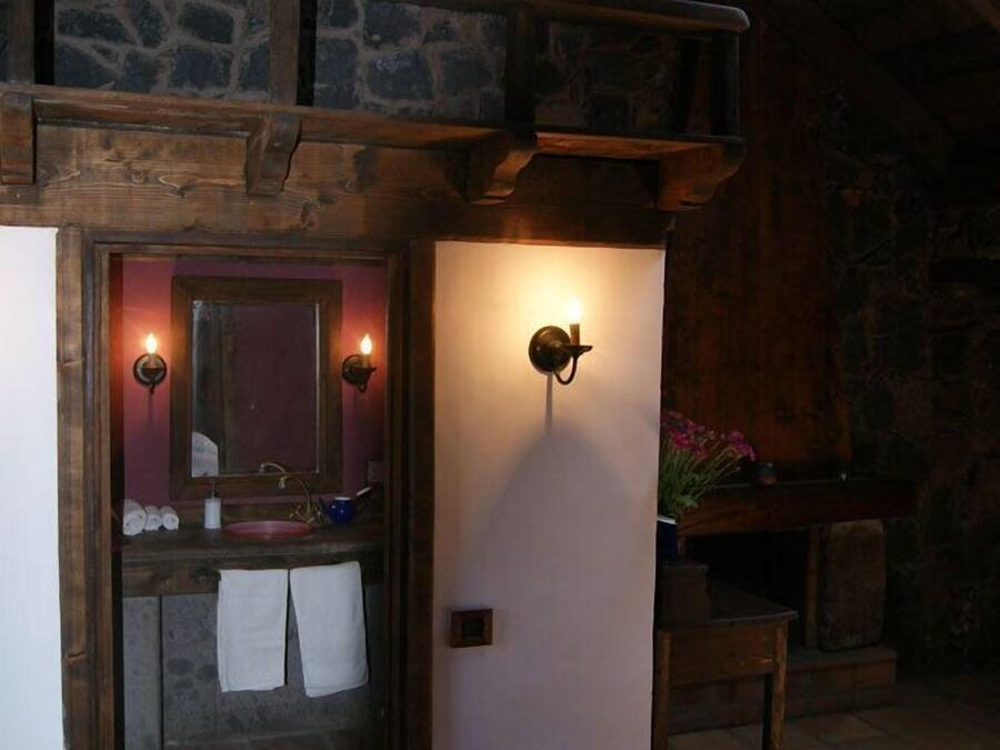 Un baño rústico con pared de piedra, vigas de madera, un lavabo con espejo y dos luces fijadas a la pared.