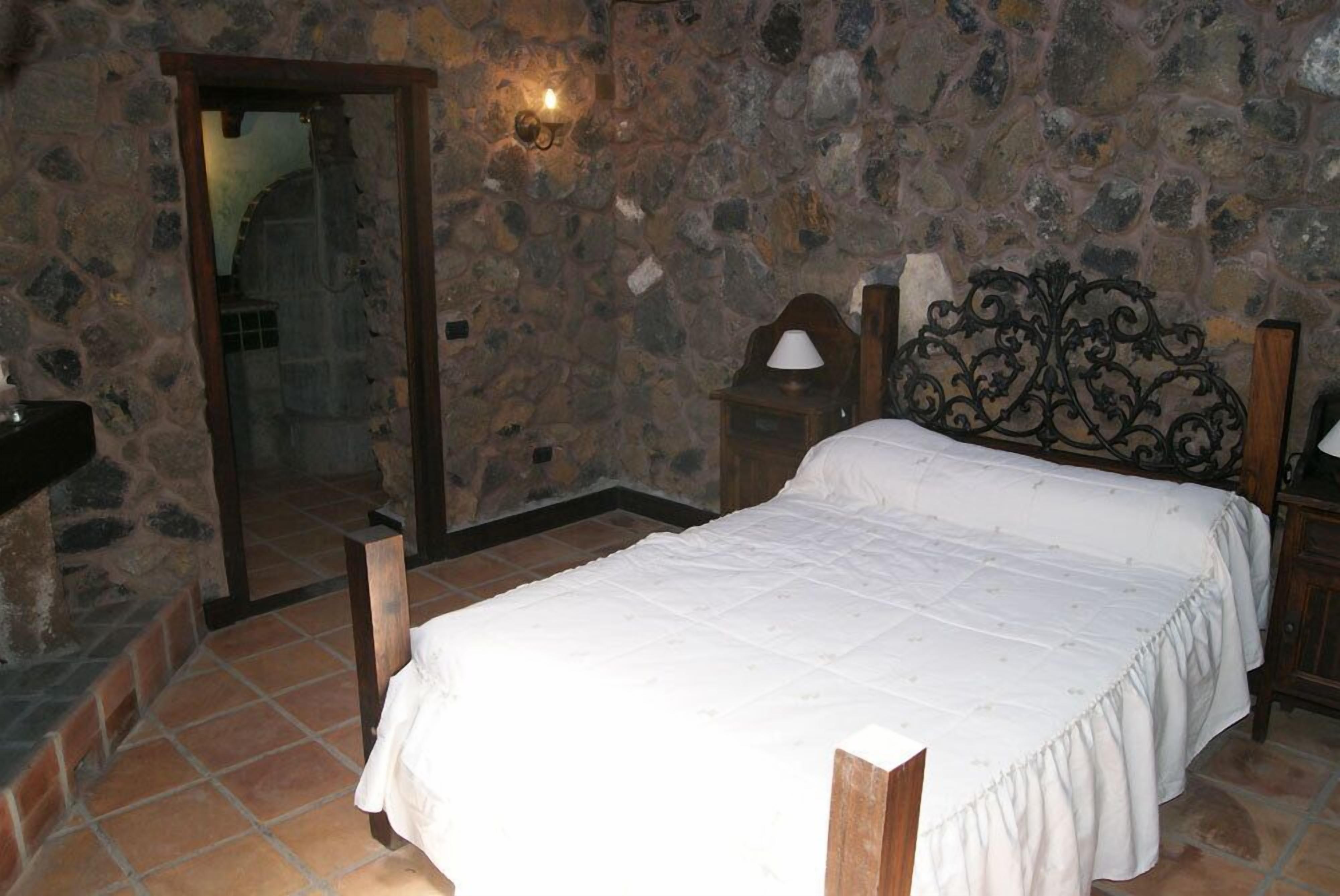 A bedroom with a stone wall, a bed with a wooden headboard, a nightstand, and a mirror.