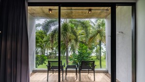 A room with a large sliding glass door leading to a balcony with a view of palm trees and a grassy area. There are two chairs and a small table on the balcony floor. - Kokotel Khao Lak Lighthouse (Takua Pa)