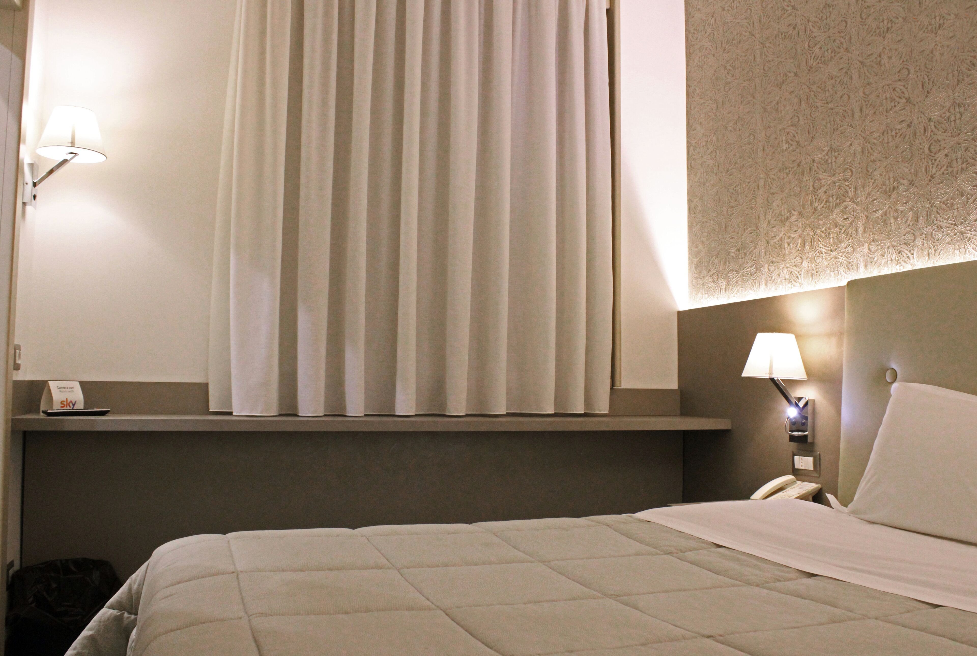 A hotel room with a bed, a window with curtains, a bedside lamp, a nightstand with a phone, and a wall-mounted light.