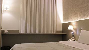 Habitación de hotel con cama, ventana con cortinas, lámpara de noche, mesita de noche con teléfono e iluminación en la pared.