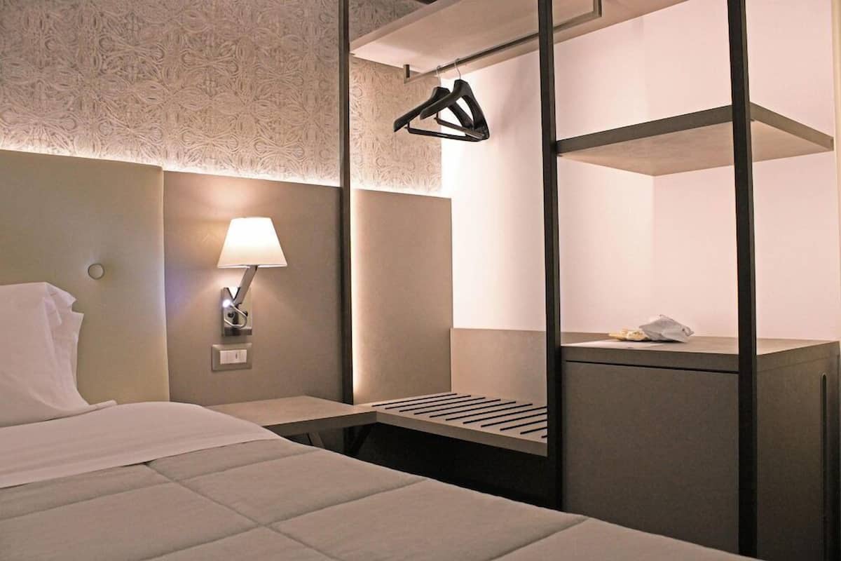 A hotel room with a bed, a bedside lamp, a nightstand, a mirror, and a hanging coat rack.