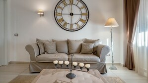 Una sala de estar con un sofá color beige, un reloj de pared grande, una lámpara de pie y una mesa de centro.