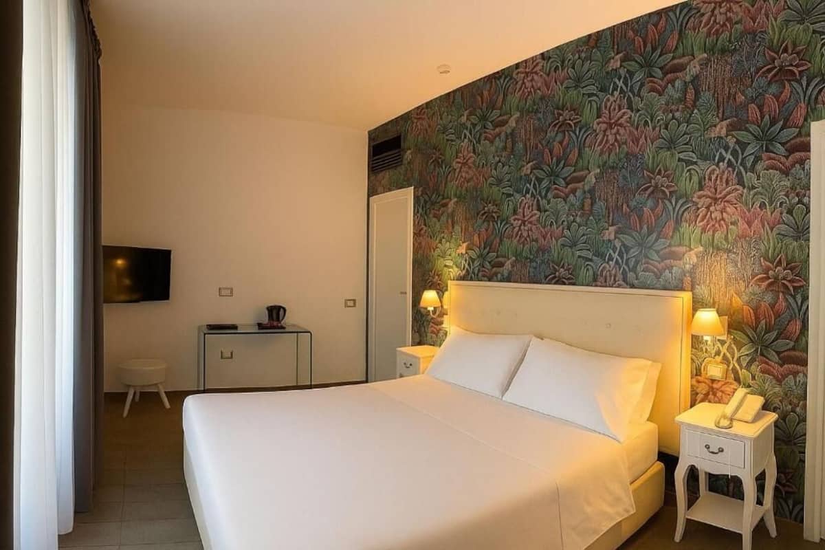 A hotel room with a large bed, two bedside tables with lamps, a flat-screen TV, a small desk, and a wall with a floral wallpaper.