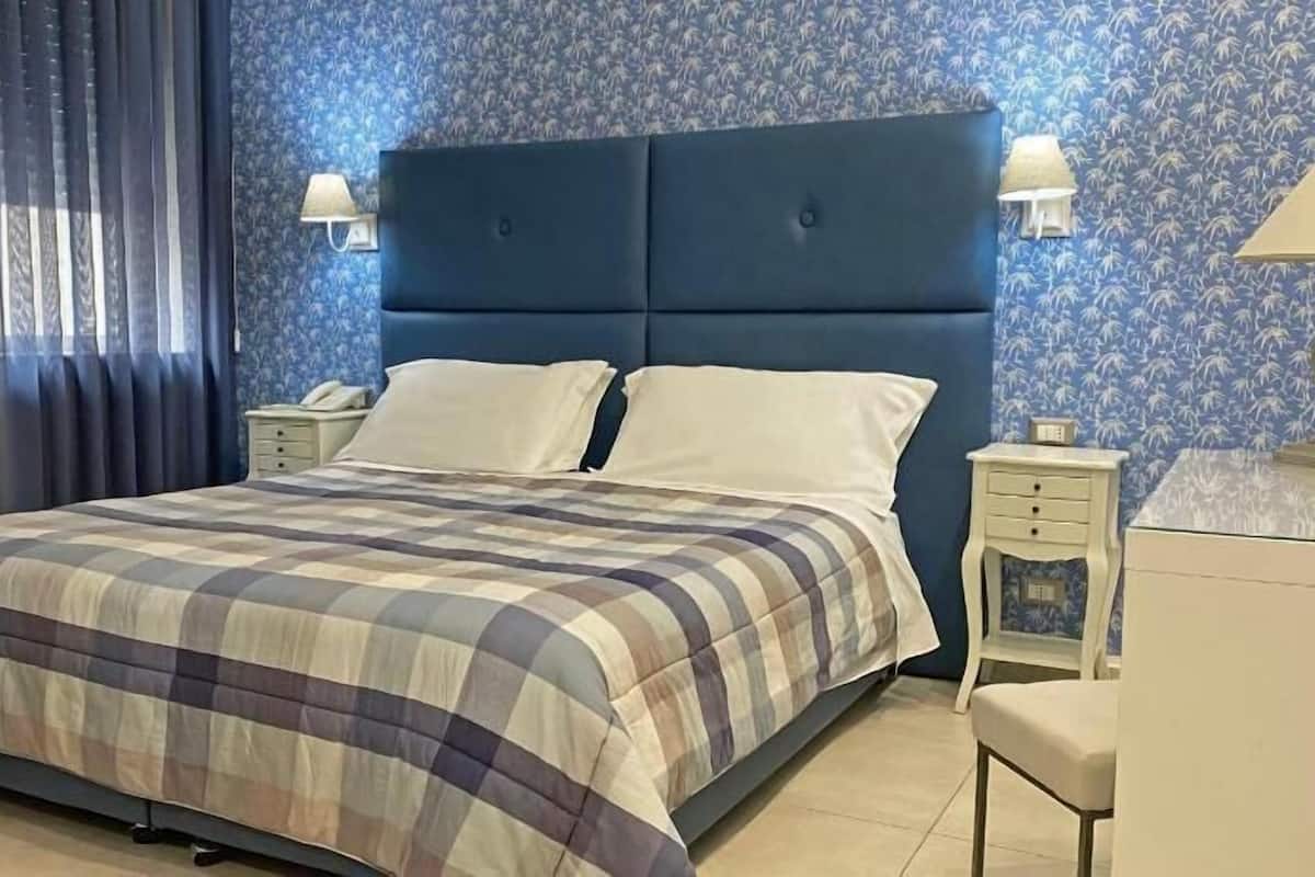 A neatly made bed with a plaid bedspread, a blue headboard, two bedside tables with lamps, and a nightstand with a lamp.