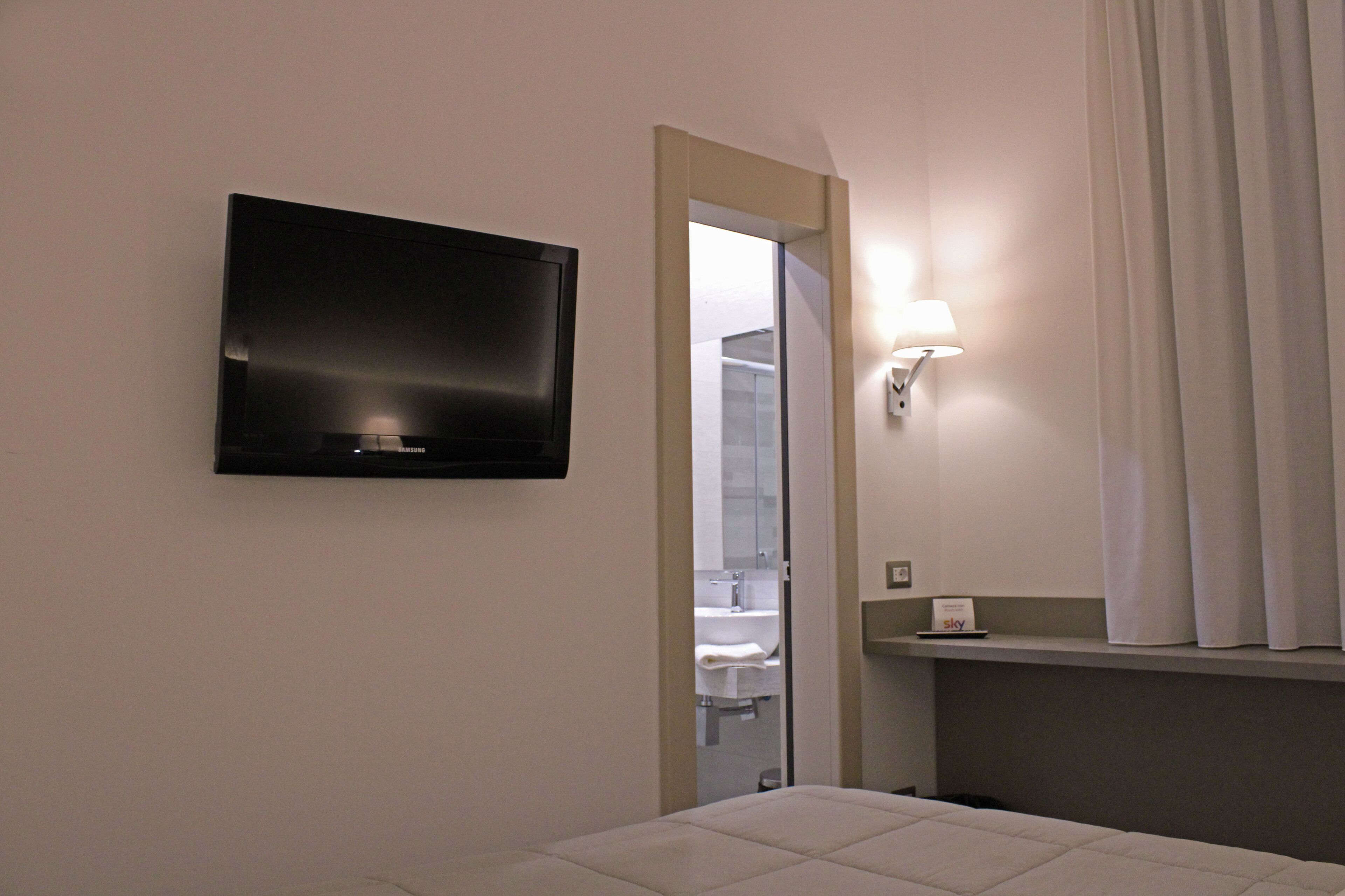 A modern hotel room with a flat-screen TV mounted on the wall, a bedside lamp, a small shelf with a 'sky' sign, and a view into the bathroom through a door.