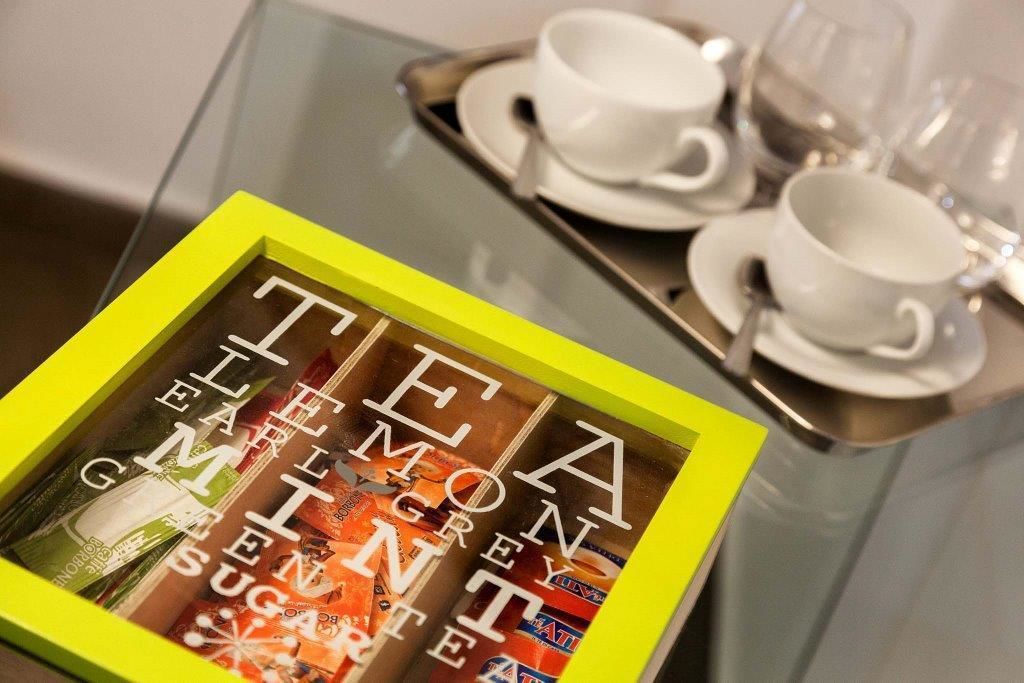 A glass table with a tray holding two white cups and saucers. In front of the table is a yellow-framed box with the text "TEA, ELLEN GIMNET, SUGAR" visible.