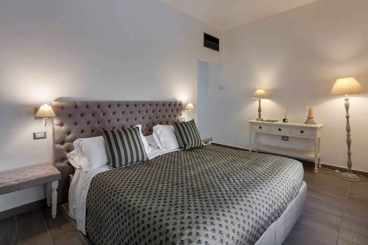 A neatly arranged bedroom with a tufted headboard, a bed with patterned bedding, bedside tables with lamps, and a nightstand.