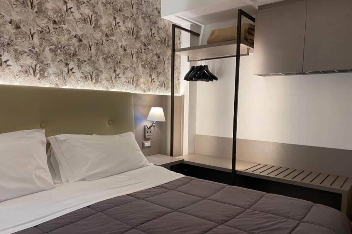 A modern hotel room with a bed, a nightstand with a lamp, a wardrobe, and a headboard with a floral pattern.