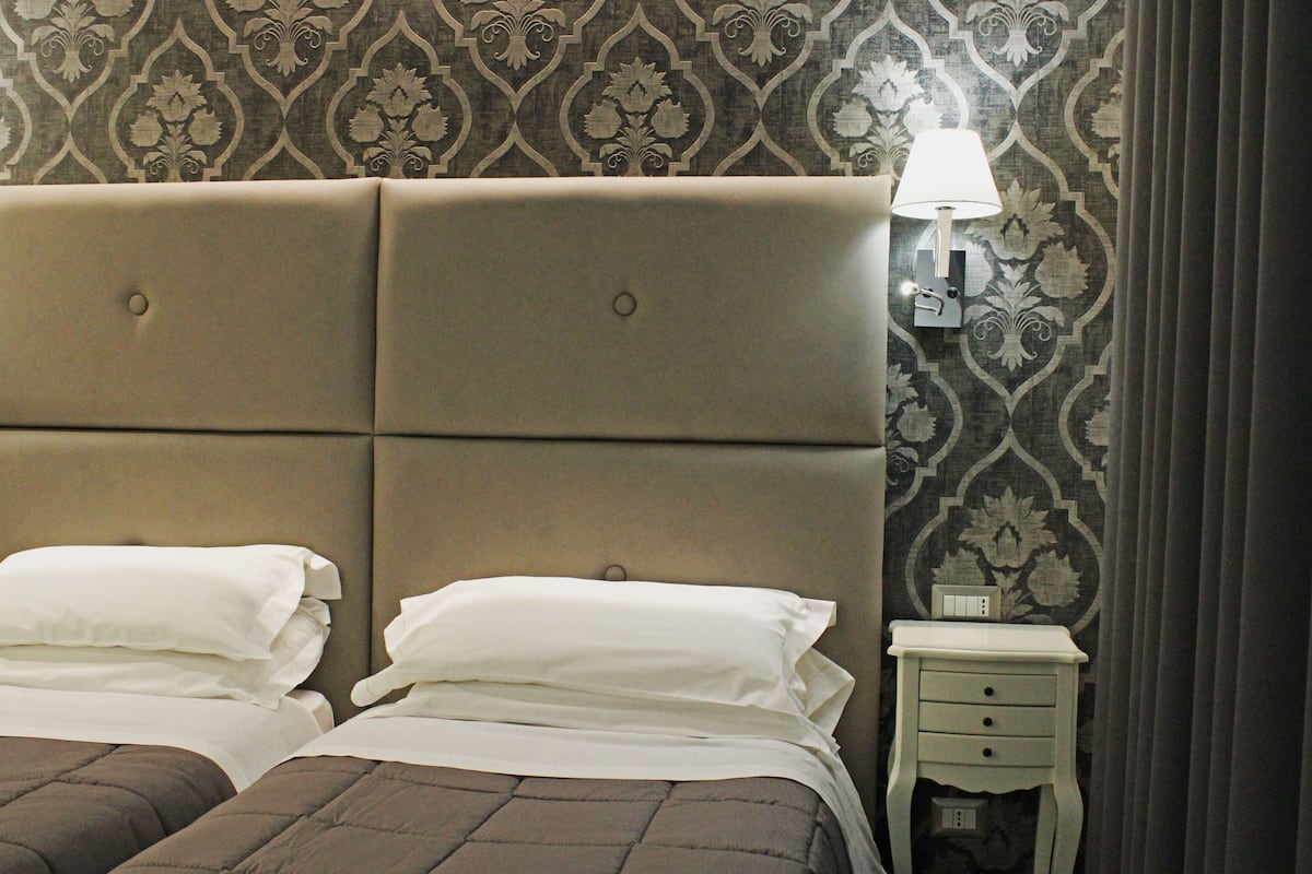 A hotel room with two beds, a nightstand with a lamp, and patterned wallpaper.