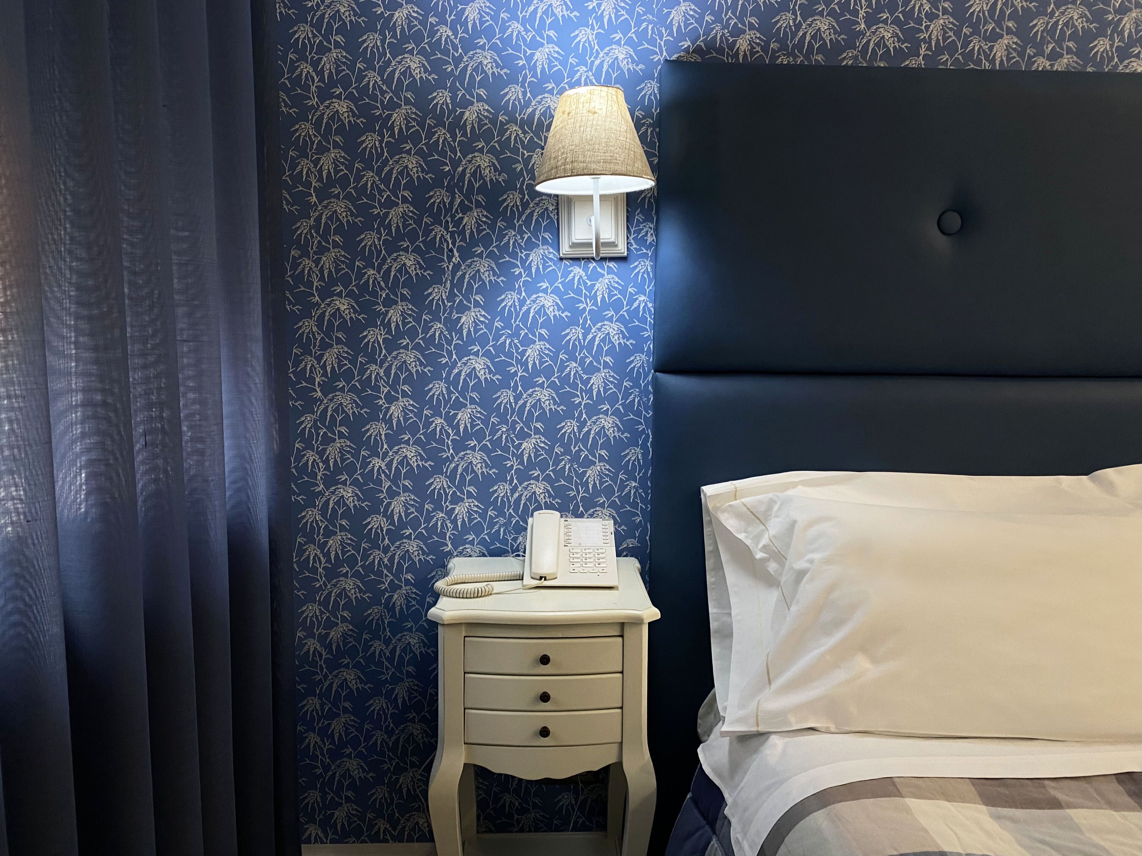 Une chambre avec un papier peint à motifs floraux bleu foncé, une lampe de chevet, une table de chevet blanche avec un téléphone, et un lit avec une literie blanche.