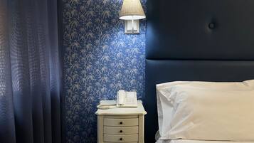 Une chambre avec un papier peint à motifs floraux bleu foncé, une lampe de chevet, une table de chevet blanche avec un téléphone, et un lit avec une literie blanche.