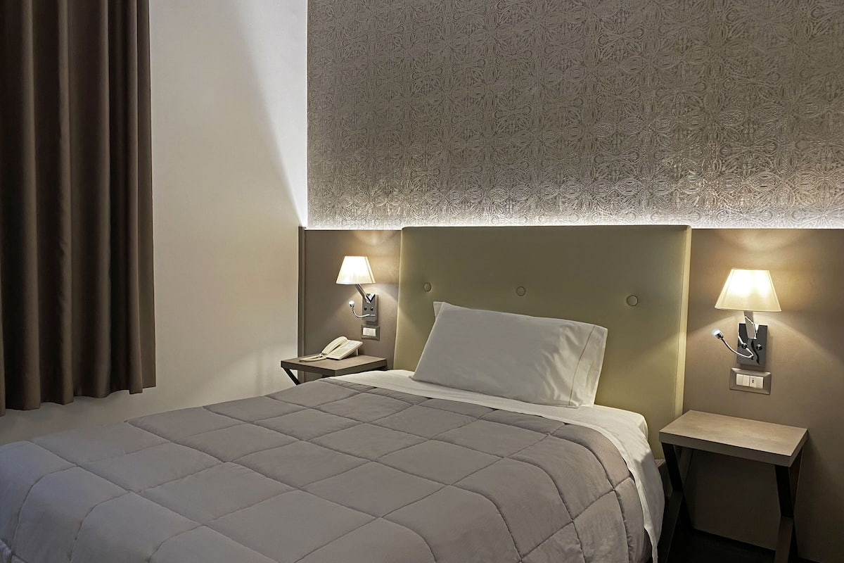 A hotel room with a bed, two bedside tables with lamps, a telephone, and a patterned headboard.