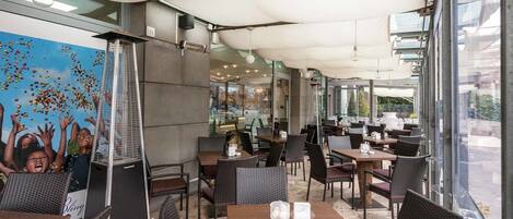 An outdoor cafe with tables and chairs, a mural on the wall, and a view of the street.