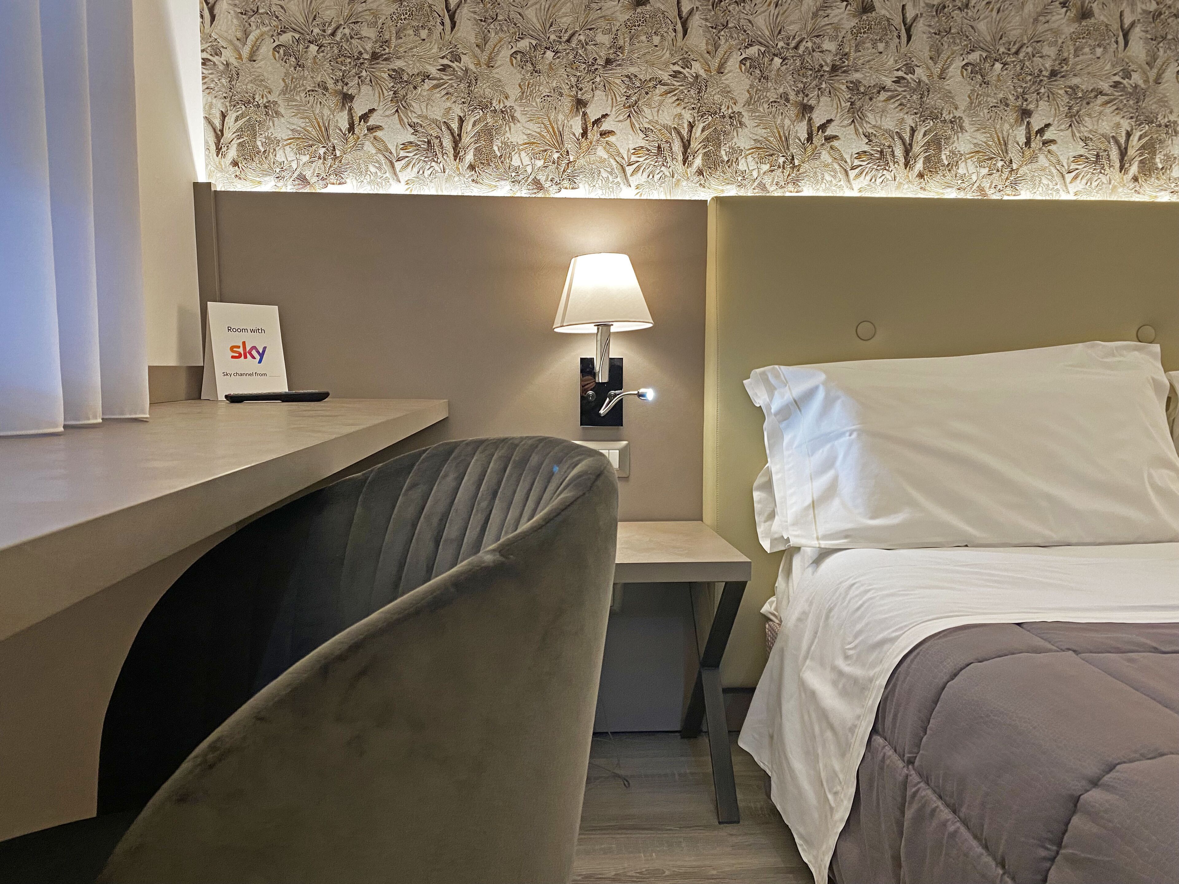 A hotel room with a bed, a desk, a chair, a lamp, and a sign indicating a Sky channel room.