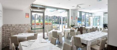 A restaurant with multiple tables set for dining, a stone wall on one side, and large windows providing a view of the outside.
