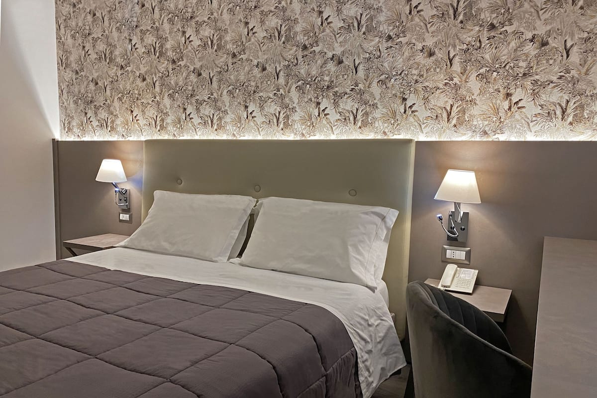A neatly made bed with a dark brown quilt, two bedside lamps, and a floral-patterned wallpaper.