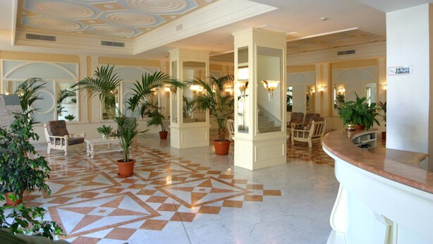 A spacious lobby with a geometric patterned floor, potted plants, and a reception desk.