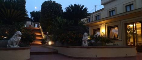 Un patio con una escalera que conduce a un edificio, rodeado de plantas y dos estatuas de leones de piedra.