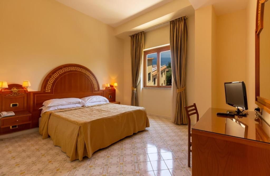 Een hotelkamer met een bed, nachtkastjes, een bureau met een computer en een raam met gordijnen.