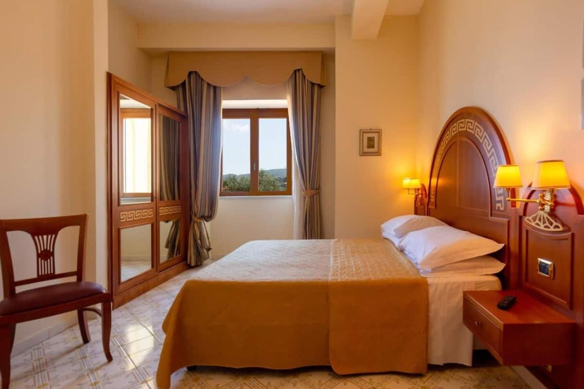 Une chambre d’hôtel avec un lit, une chaise, une table de chevet avec des lampes et une vue sur l’extérieur par une fenêtre.