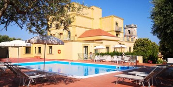 Una piscina con lettini e ombrelloni, un edificio con tetto in tegole rosse e un campanile.