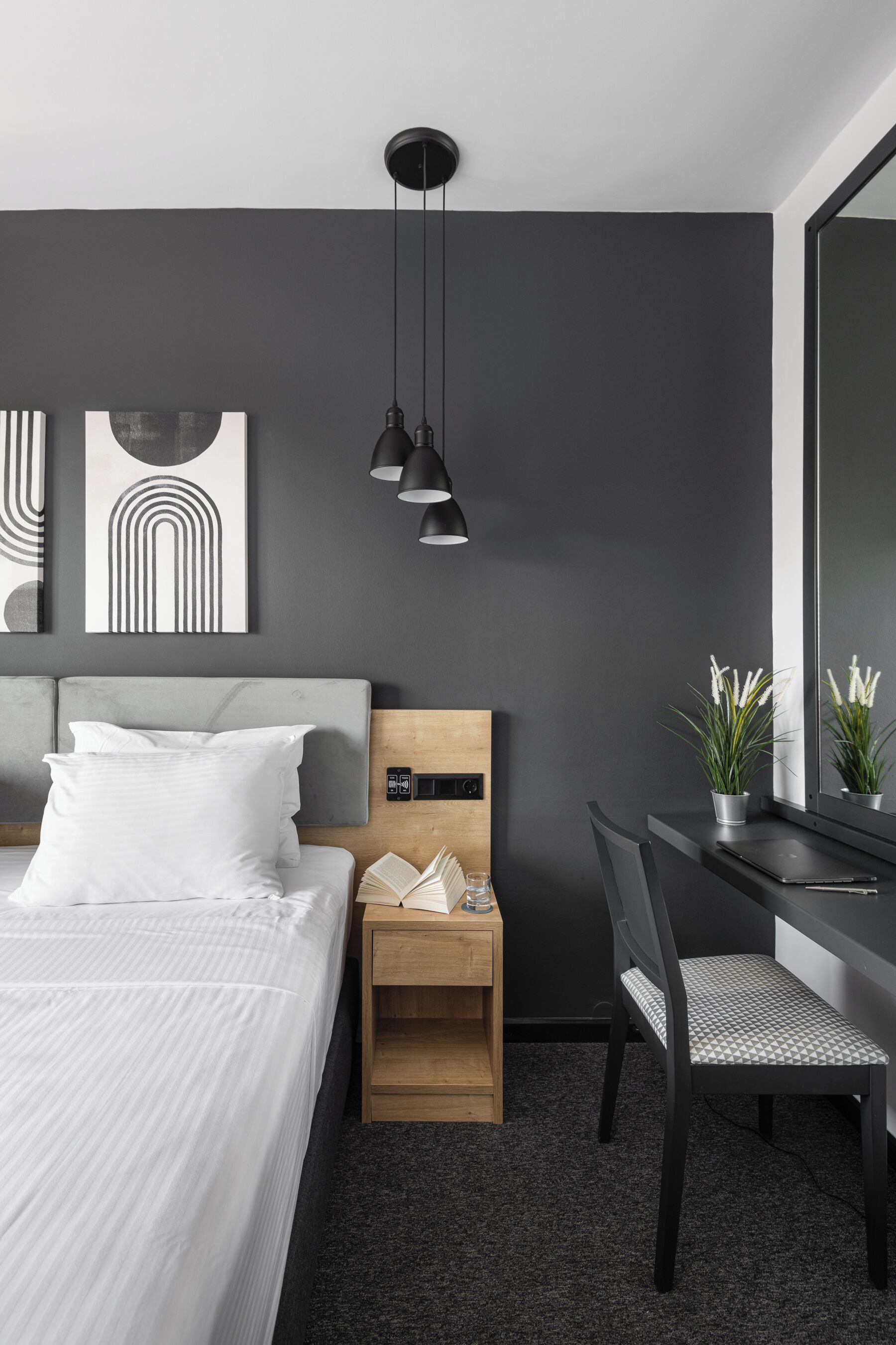 A modern hotel room with a large bed, a desk with a laptop, a chair, a nightstand, and a mirror.