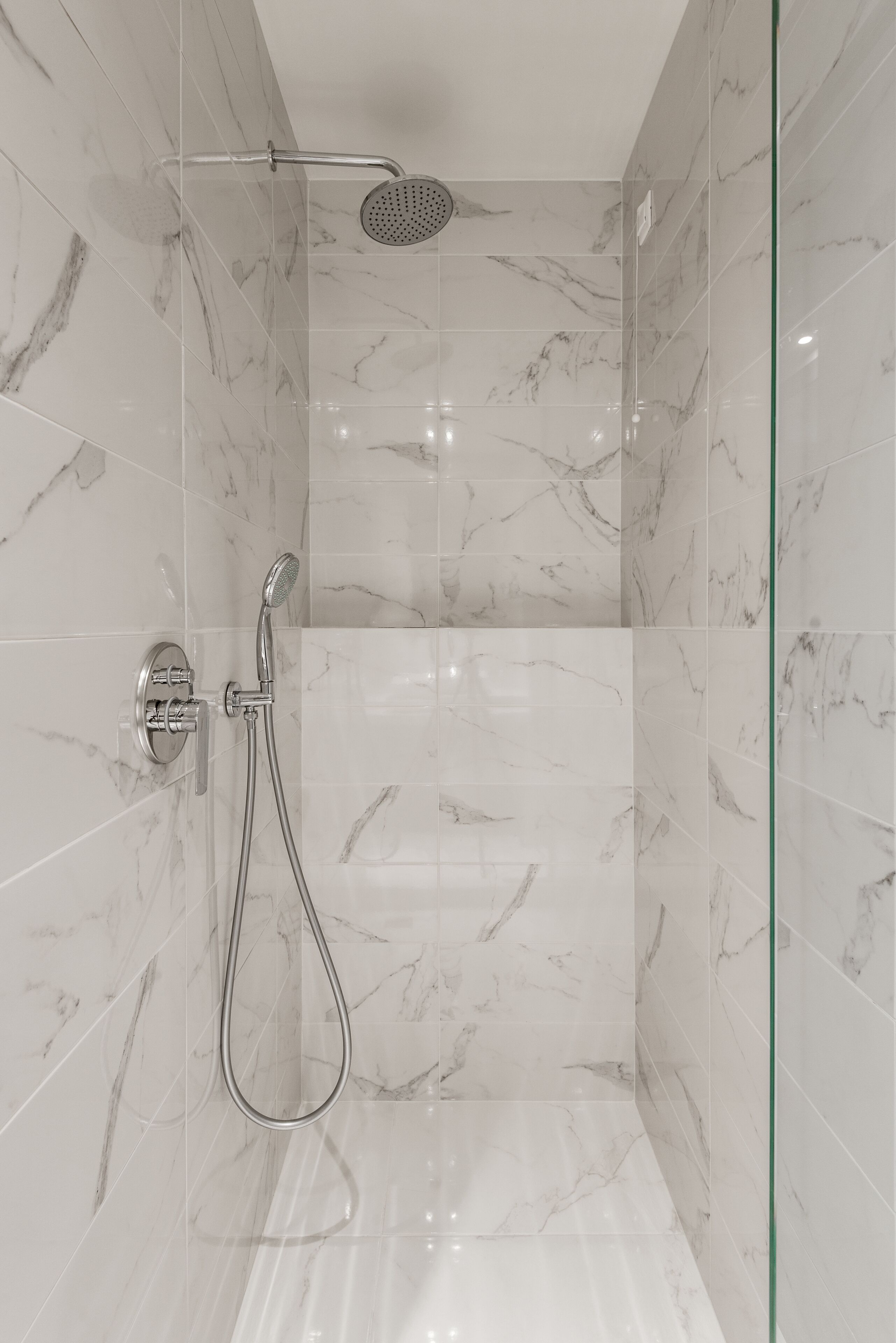 A modern bathroom with marble tiles, a shower with a handheld showerhead, and a glass-enclosed shower area.