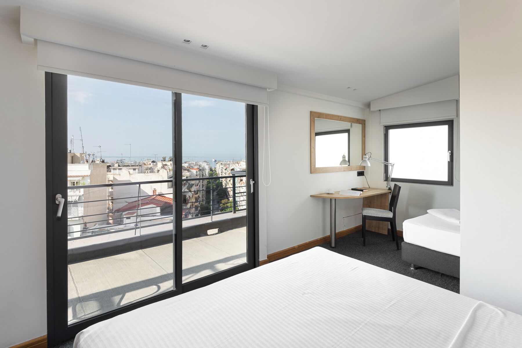 Habitación de hotel con un gran ventanal corredero, un escritorio con silla, una cama y ventana con vistas a la ciudad.