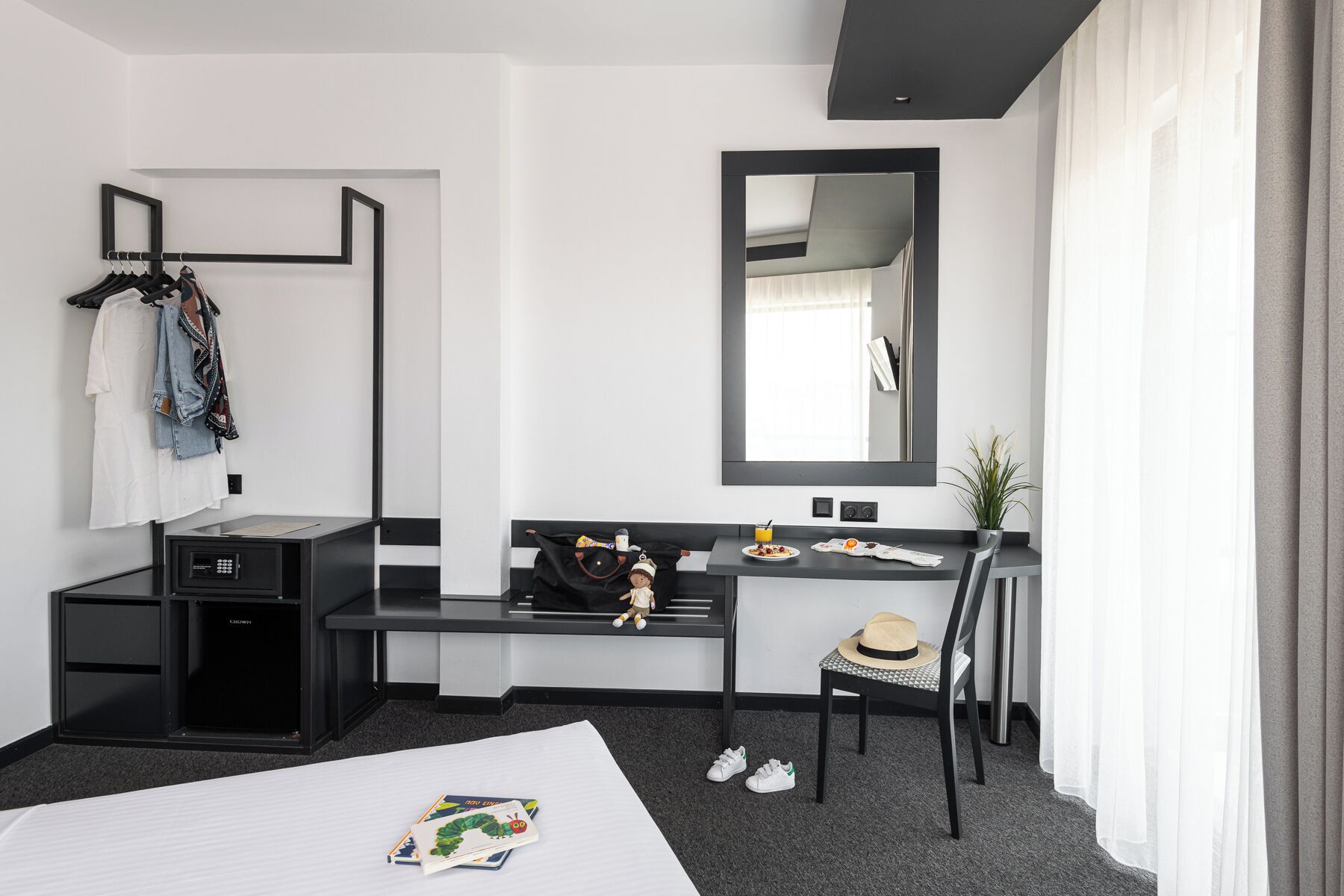 A hotel room with a bed, a desk with a chair, a wardrobe, a mirror, and a small table with a plant.